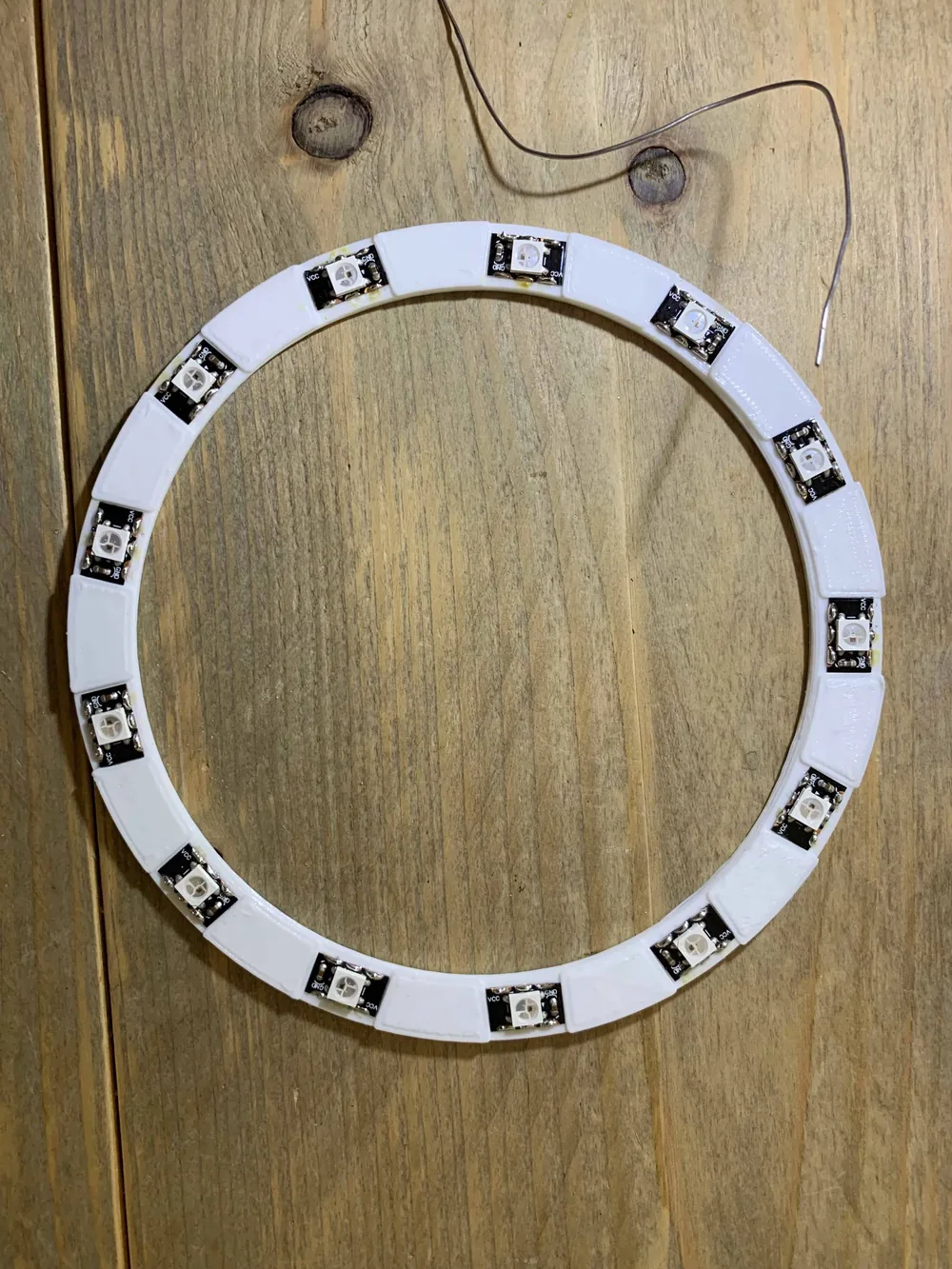 A white circular circuit board with sixteen NeoPixel LEDs and a thin cable, lying on a wooden table